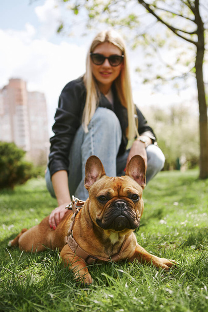 Beautiful blonde woman walking the dog in sunny spring park. Cute brown French bulldog lying on grass and looking in camera - Foto, immagini
