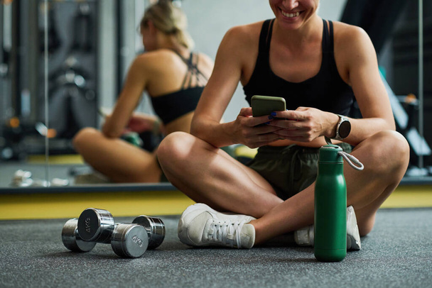 Κοντινό πλάνο της νεαρής γυναίκας με αθλητικά είδη που κάθεται στο πάτωμα και χρησιμοποιεί το κινητό τηλέφωνο μετά την προπόνηση στο γυμναστήριο - Φωτογραφία, εικόνα