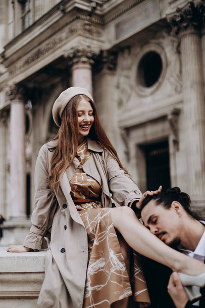 A couple in love walks along the streets of Paris, a guy in a coat and a suit, a girl in a dress with a beret and a trench coat in autumn - Foto, Imagen