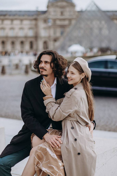 Ένα ερωτευμένο ζευγάρι περπατά στους δρόμους του Παρισιού, ένας τύπος με παλτό και κοστούμι, ένα κορίτσι με φόρεμα με μπερέ και καμπαρντίνα το φθινόπωρο. - Φωτογραφία, εικόνα
