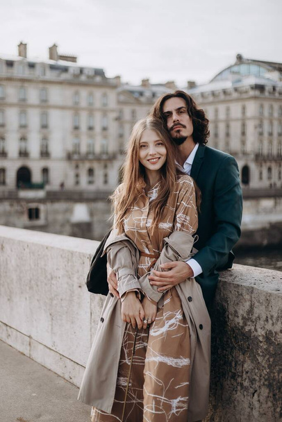 A couple in love walks along the streets of Paris, a guy in a coat and a suit, a girl in a dress with a beret and a trench coat in autumn - Фото, изображение