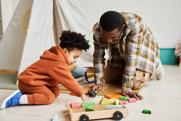 Full length portret van zorgzame zwarte vader spelen met zoon op de vloer in gezellige kinderkamer, vaderschap en familie concept - Foto, afbeelding