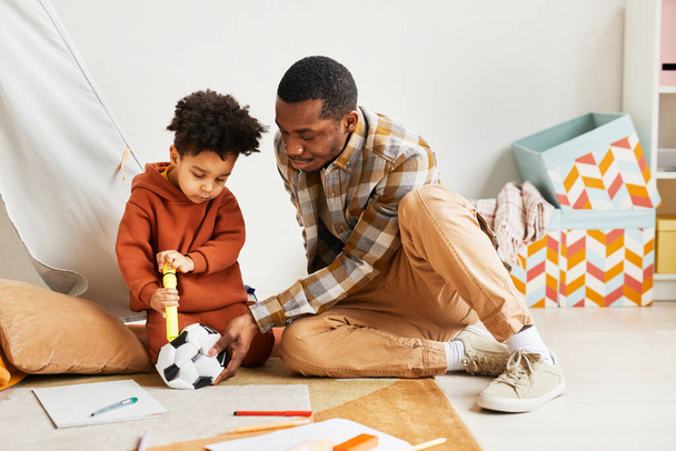 Портрет заботливого чернокожего отца в полный рост, играющего с маленьким сыном в закрытом помещении и прищипывающего детский футбольный мяч, копирующее пространство - Фото, изображение