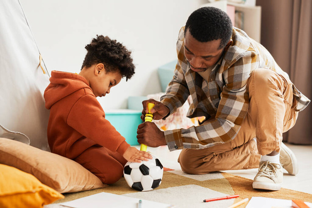 Portrait latéral de père noir attentionné jouant avec un tout-petit fils à l'intérieur et pompant le ballon de football pour enfants - Photo, image