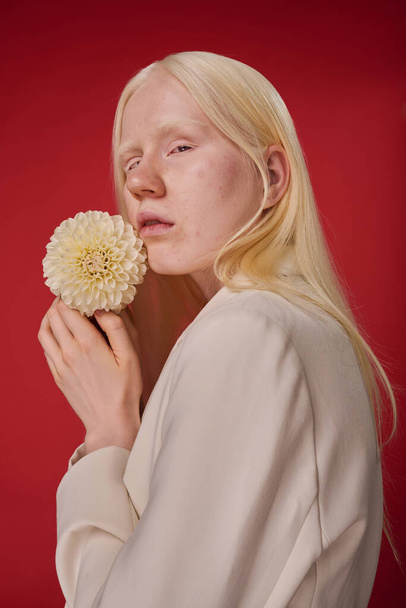 Portré gyönyörű albínó lány fehér haj gazdaság virág mellett arca elszigetelt piros háttér - Fotó, kép