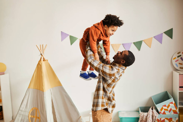 Minimal side view portrait of loving black father playing with toddler son at home and holding him up in air, copy space - Foto, immagini