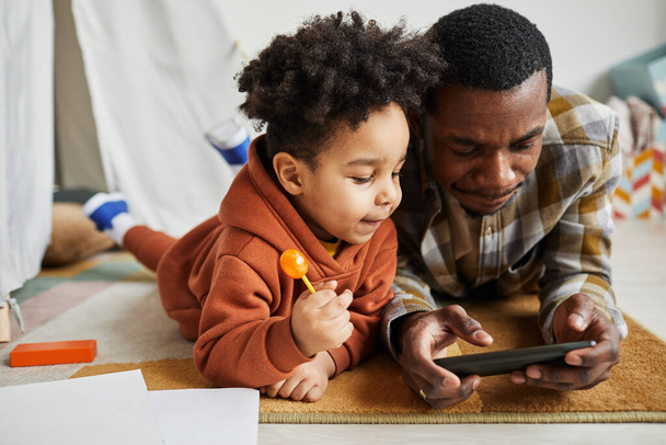 Muotokuva musta isä lapsi poika makaa lattialla yhdessä ja katsella sarjakuvia älypuhelimella leikkihuoneessa - Valokuva, kuva