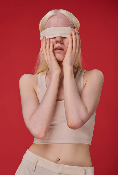 Portrait of young slim woman with blindfolded eyes standing against red background - Fotoğraf, Görsel