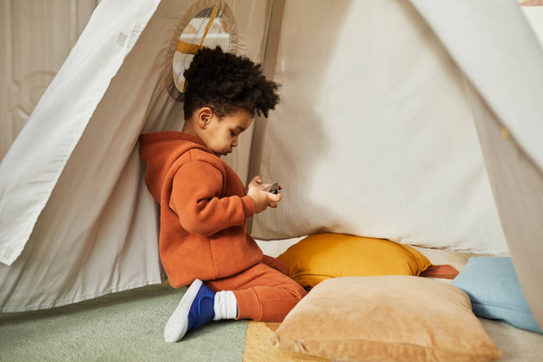 Küçük Afro-Amerikan çocuğun oyun çadırında akıllı telefon kullandığı ve modaya uygun kıyafetler giydiği minimal yan görüntü portresi. Alfa jenerasyonu, kopyalama alanı. - Fotoğraf, Görsel