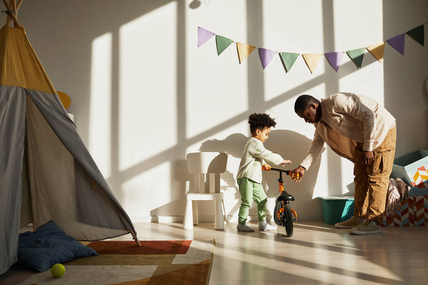 Retrato mínimo de padre negro amoroso enseñando a niño pequeño a montar en bicicleta de equilibrio en casa a la luz del sol, espacio de copia - Foto, imagen