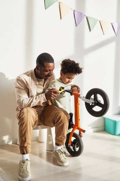 Pionowy portret kochającego czarnego ojca uczy małego chłopca jeździć na rowerze w domu w świetle słonecznym - Zdjęcie, obraz