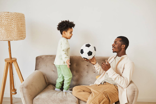 Asgari yandan bakan mutlu zenci baba portresi. Evde yeni yürümeye başlayan oğluyla oynuyor ve elinde futbol topu tutuyor. - Fotoğraf, Görsel
