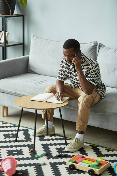 Vertical portrait of young black man working from home and talking on smartphone in grey interior - Photo, Image