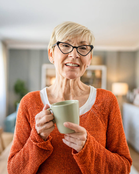 Primer plano retrato de una mujer mayor con el pelo corto sonrisa feliz emoción positiva espacio de copia de pie en casa gafas de desgaste interior tienen una taza de café rutina diaria por la mañana - Foto, imagen