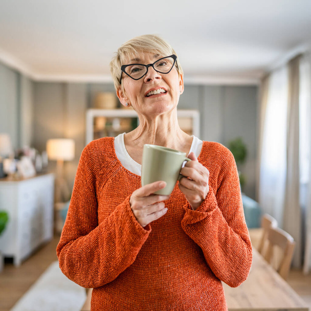 Κοντινό πλάνο πορτρέτο μιας ηλικιωμένης γυναίκας με κοντά μαλλιά χαρούμενο χαμόγελο θετική συγκίνηση αντίγραφο χώρο στέκεται στο σπίτι εσωτερική φορούν γυαλιά έχουν ένα φλιτζάνι καφέ καθημερινή ρουτίνα το πρωί - Φωτογραφία, εικόνα