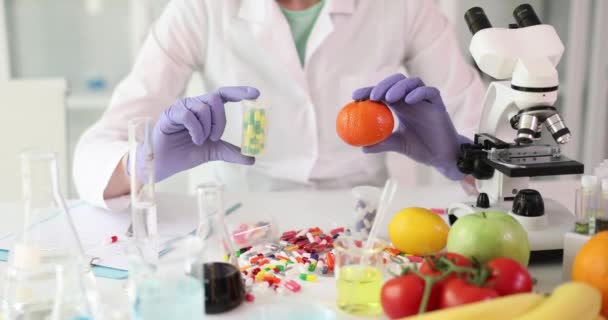 Científico sosteniendo mandarina o naranja y pastillas en el laboratorio. Vitaminas sintéticas y daños para la salud humana - Imágenes, Vídeo