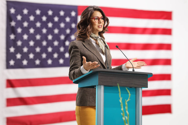 Young woman on a pedestal giving a speech and gesturing with hands with the USA flag in the background - Фото, изображение