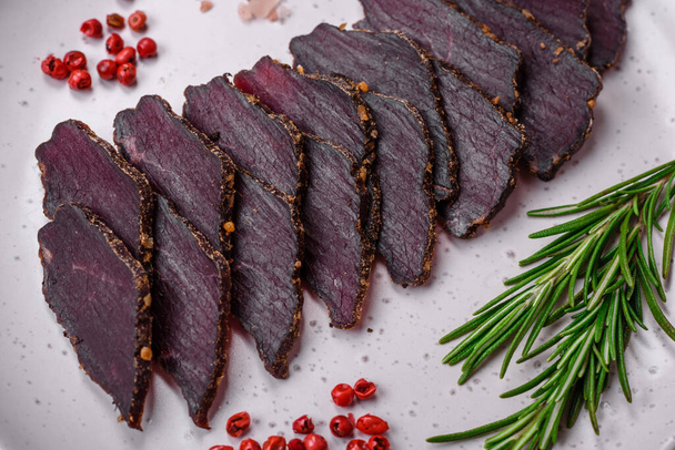 Delicious dried smoked beef or horse meat jerky with spices and salt on a dark concrete background - Photo, Image