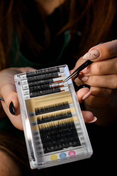 close-up on a plastic box with artificial eyelashes of different sizes in a female hand. Close-up tool for eyelash extension. Cosmetician takes eyelashes with tweezers - 写真・画像