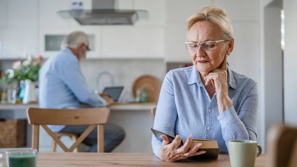 Jedna žena zralý senior kavkazský žena důchodce moderní babička číst knihu doma nosit brýle, zatímco její manžel stojí za rodinným životem aktivní senior skuteční lidé kopírovat prostor - Fotografie, Obrázek