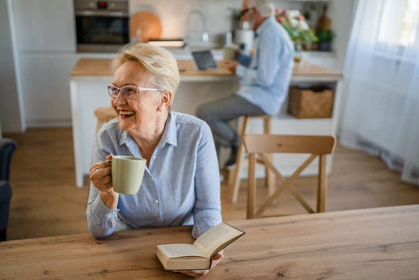 One woman mature senior caucasian female pensioner modern grandmother read book at home wear eyeglasses while her husband stand behind family life active senior real people copy space - Fotoğraf, Görsel