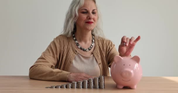 Happy middle aged elderly woman puts coin in piggy bank and saves money for future. Retired woman making investments and family savings - Footage, Video