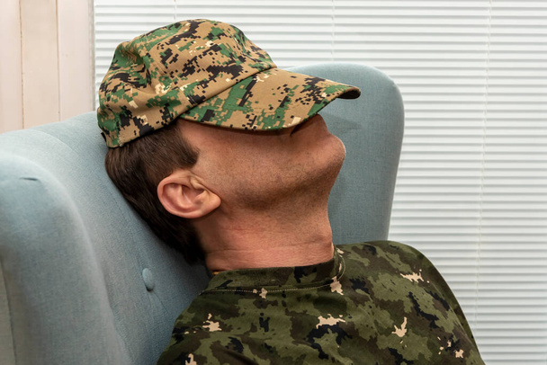 Vanhempi sotilas 45-50-vuotias peitti kasvonsa armeijan lakilla, nukkuen nojatuolissa. Käsite: mielenterveyshäiriöt sotilashenkilöstössä, psykoterapeutin hoito. - Valokuva, kuva
