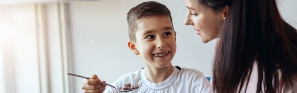 Retrato, pequeno menino adolescente comendo cereais com leite em kichen e olhando para a mãe. Tomar café da manhã. - Foto, Imagem