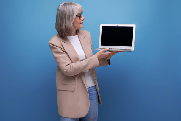60s γυναίκα στο σακάκι δείχνει mockup οθόνη laptop σε φωτεινό φόντο με αντίγραφο χώρο. - Φωτογραφία, εικόνα