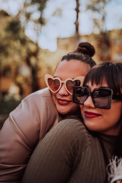 Two young women embrace each other emotionally outside on a sunny day while wearing sunglasses.  - Foto, afbeelding