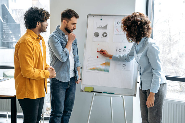 Focused busy team of multiracial financial analysts, economic experts, stand in a modern office near a whiteboard, concentrated discuss financial strategy, plan, perspective, share ideas and thoughts - Φωτογραφία, εικόνα