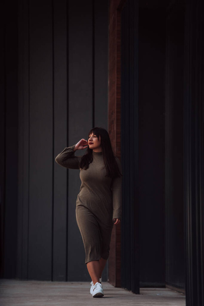 Una donna fiduciosa ed elegante plus-size a piedi da un edificio moderno in un abito verde oliva, che irradia bellezza, emancipazione e amore per se stessi - Foto, immagini