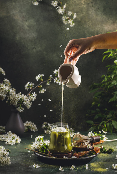Женская рука наливает самодельную сметану из маленькой банки в стакан чаем маття. Темный фон с весенними вишневыми бочками и традиционными блюдами для этого чая - Фото, изображение
