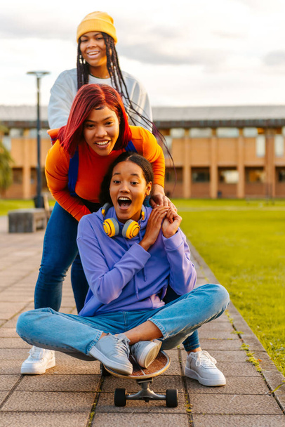 Multiracial (Latina and black) group of three teenage friends having fun together on a skateboard outside of school. Unity and friendship. classmates - 写真・画像