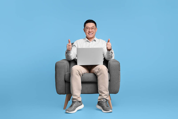 Happy man with laptop showing thumb up gesture while sitting in armchair on light blue background - Foto, immagini