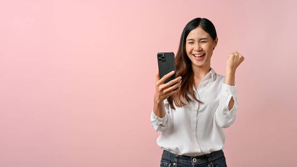 Young happy Asian woman celebrate with herself while looking at smartphone in pink isolated background.  - Photo, Image