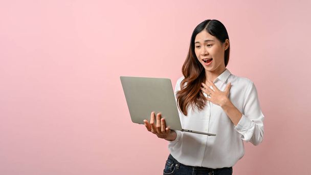 Entzückende junge Asiatin, die überrascht ist, als sie auf ihren Laptop blickt und vor rosa Hintergrund steht.  - Foto, Bild