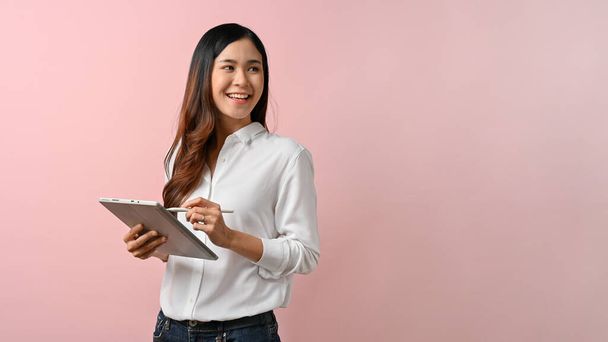 Image of young Asian woman holding digital tablet while standing with pink isolated background. - Photo, Image