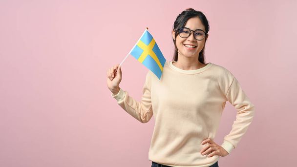 Nuori aasialainen tyttö näyttää Ruotsin lipun vaaleanpunainen eristetty tausta, koulutus käsite. opiskella ulkomailla käsite. - Valokuva, kuva