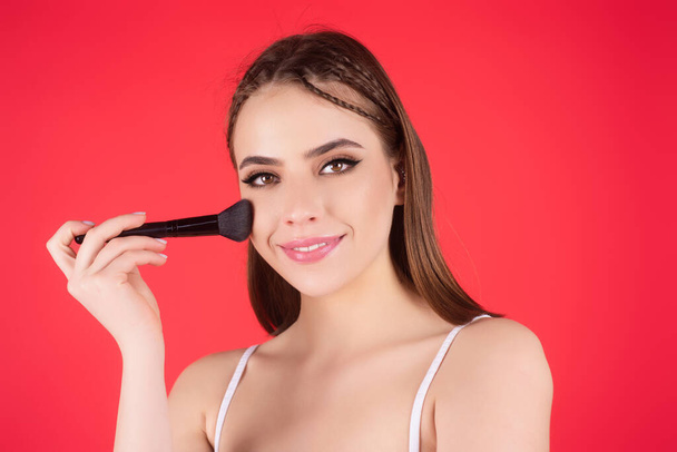 Girl hold blush blusher apply powder visage isolated over studio background. Young woman powdering cheeks. Makeup brush. Female model gets blush powder on the cheekbones - Photo, Image