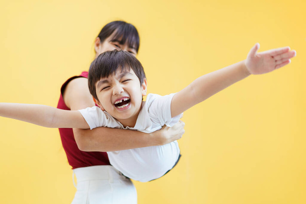 Happy cheerful Asian woman and a little young boy playing together, a woman piggyback or carrying a little boy on her back. Woman and boy portrait on yellow pastel background. - Photo, Image