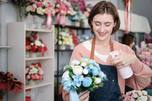 Portrait of one young Caucasian female florist worker in apron water spraying bunch of blossom in bright and beautiful flower shop store with smile, small business occupation, happy SME entrepreneur.  - Foto, Bild