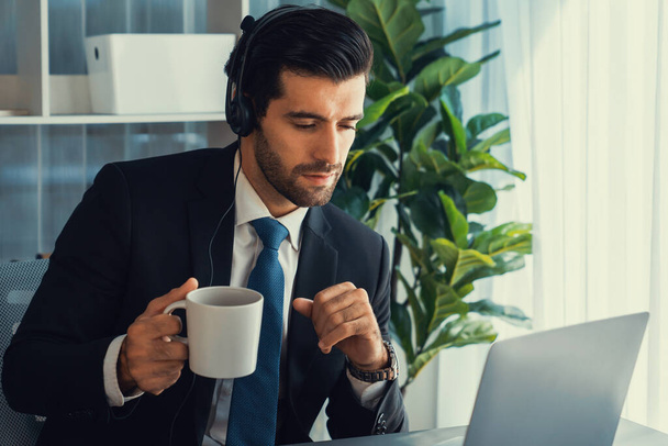 Manager biura operatora call center siedzi na biurku z kawą podczas pracy na laptopie. Pracownik biurowy noszący słuchawki i czarny garnitur pracujący nad obsługą klienta lub telemarketingiem. żarliwy - Zdjęcie, obraz
