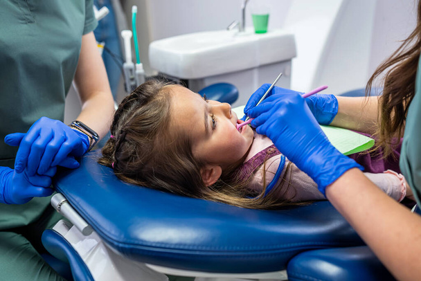 een klein meisje wordt behandeld voor kleine tanden in een tandartsstoel door een gekwalificeerde kinderarts. Het concept van tandheelkundige behandeling bij peuters - Foto, afbeelding