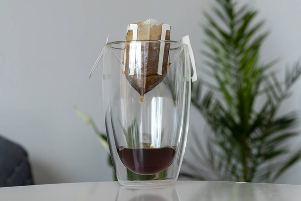 Der Prozess des Aufbrühens eines morgendlichen Kaffeetropfens mit einem Filterbeutel in einer durchsichtigen Tasse mit doppeltem Boden. - Foto, Bild