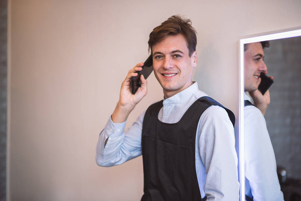 Retrato sonriente joven adulto caucásico masculino peluquero profesional usando teléfono inteligente para hablar con el cliente para la reserva de peluquería, Pequeña empresa en la ciudad. - Foto, Imagen