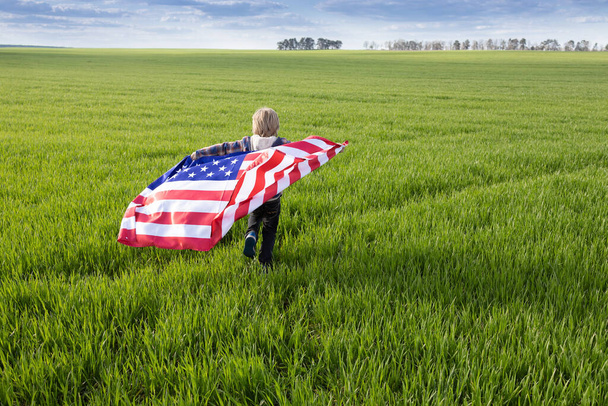 Día de la Ndependencia. Símbolo nacional de libertad e independencia de los Estados Unidos de América. niño sostiene la bandera americana detrás de la espalda y corre a través del prado verde. Orgullo de ser americano - Foto, Imagen