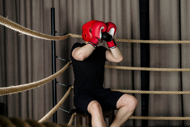 El tipo grande se sienta en la esquina de un ring de boxeo, vistiendo casco rojo y guantes y bebiendo de una botella deportiva - Foto, imagen