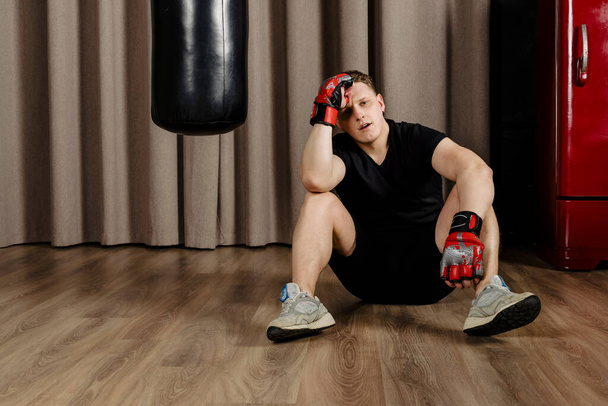 Portret zmęczonego boksera siedzi na podłodze i robi sobie przerwę, po treningu z workiem treningowym w przytulnym beżowym studio. - Zdjęcie, obraz