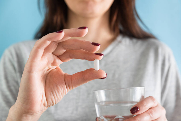 Kobieta przyjmująca białą tabletkę leku statynowego w leczeniu wysokiego poziomu cholesterolu lub środków przeciwbólowych popijając szklanką wody na niebieskim tle. Przyjmowanie leków, opieki zdrowotnej, apteki i koncepcji leczenia. Skupienie selektywne - Zdjęcie, obraz